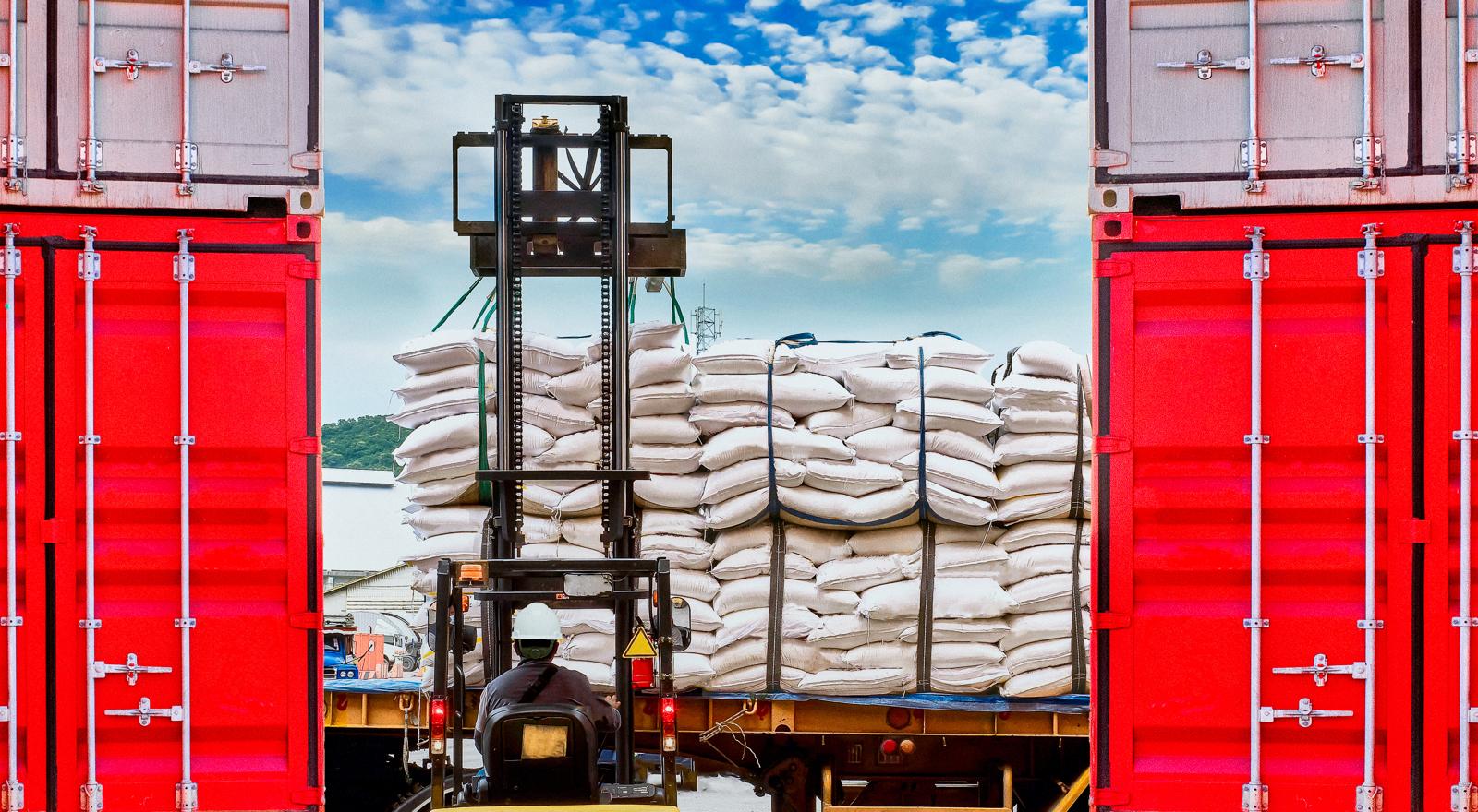 A forklift operator lifts white sugar bags onto a truck from containers